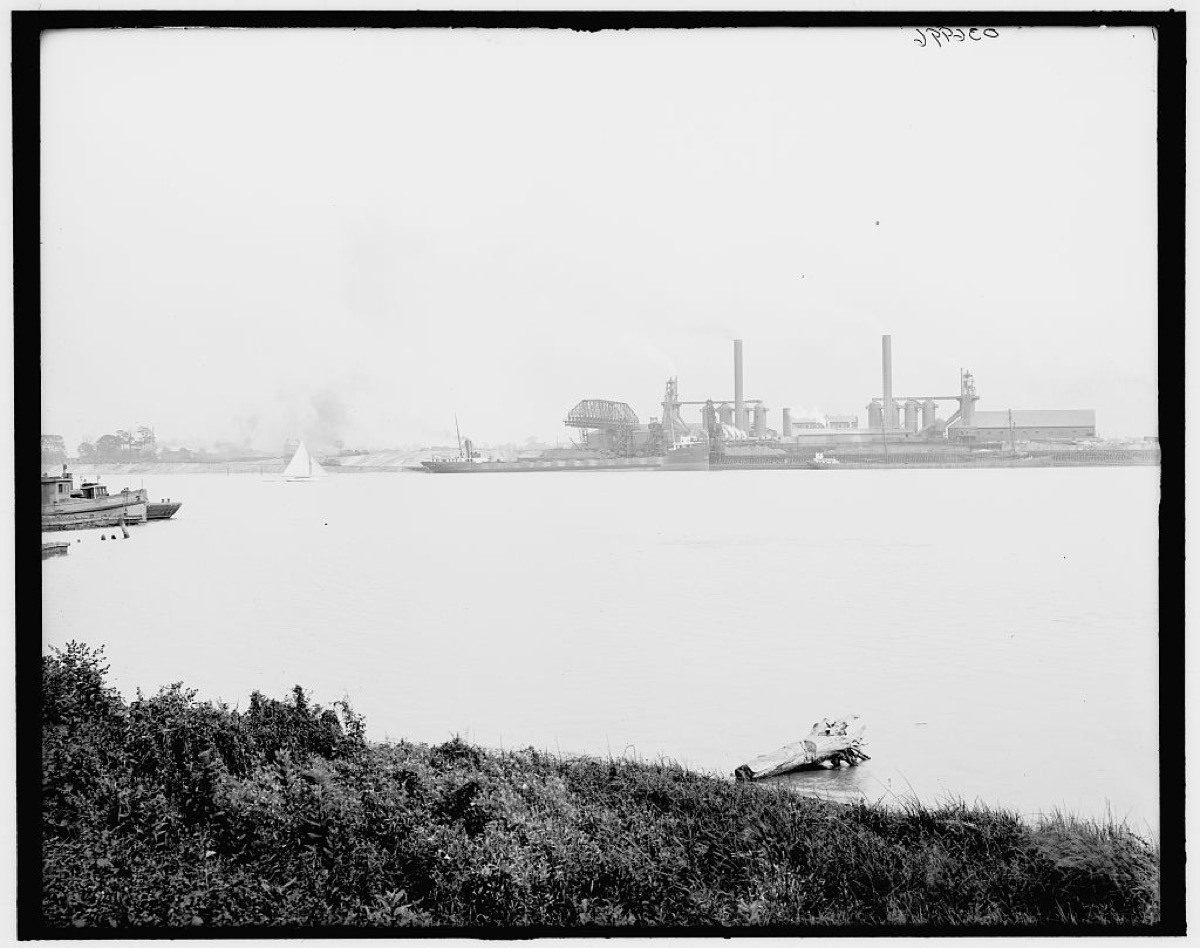 Smoke and steel on the Maumee: blast furnaces on the west bank at Toledo, 1905, a wrecked hull rotting in the shallows below. The river the whole valley was built to use runs still in front of the works that used it